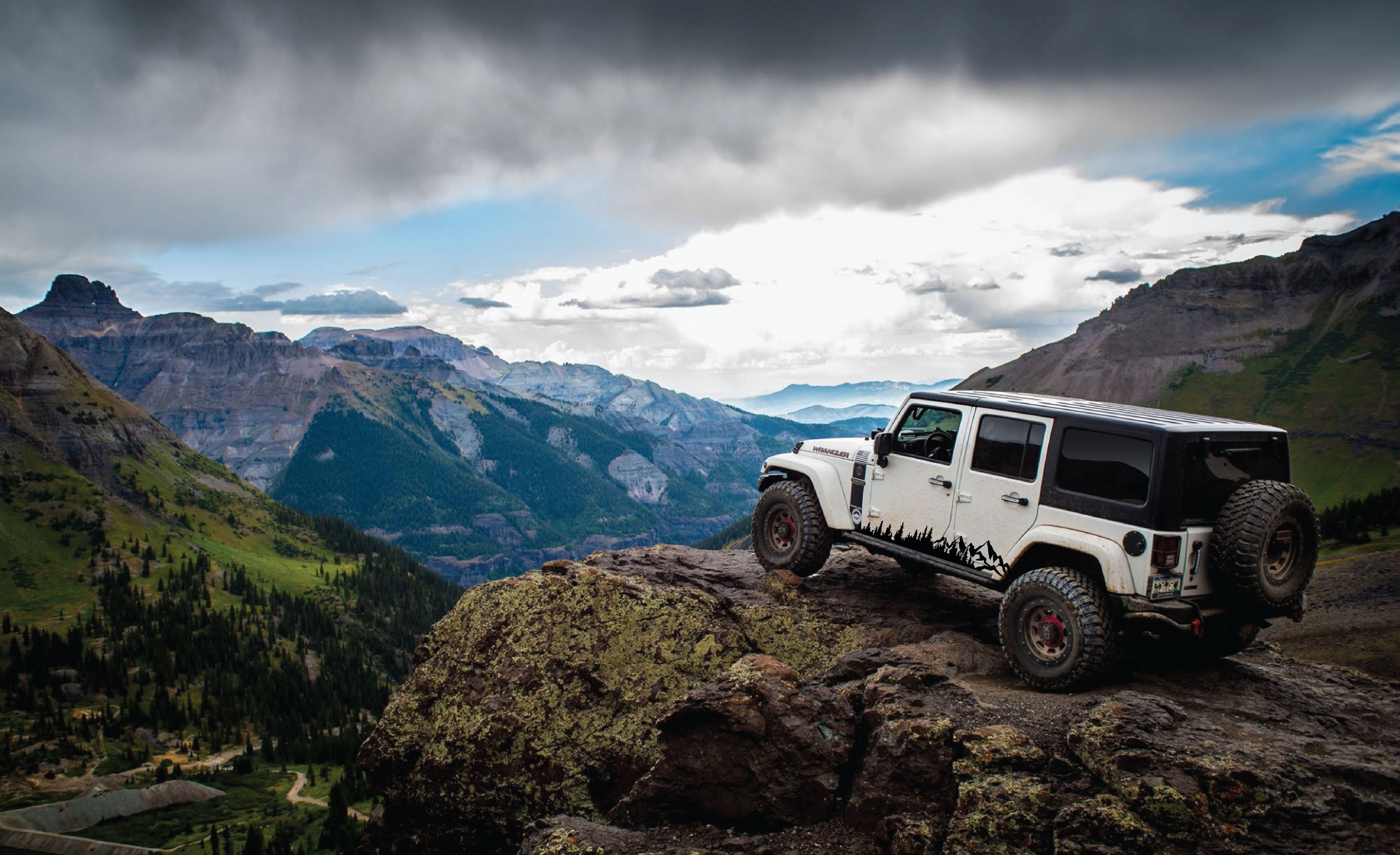 White Jeep Wrangler parked on a cliff overlooking the Rocky Mountains. Matte black vehicle mountain landscape decals installed on the bottom sides of the jeep. 